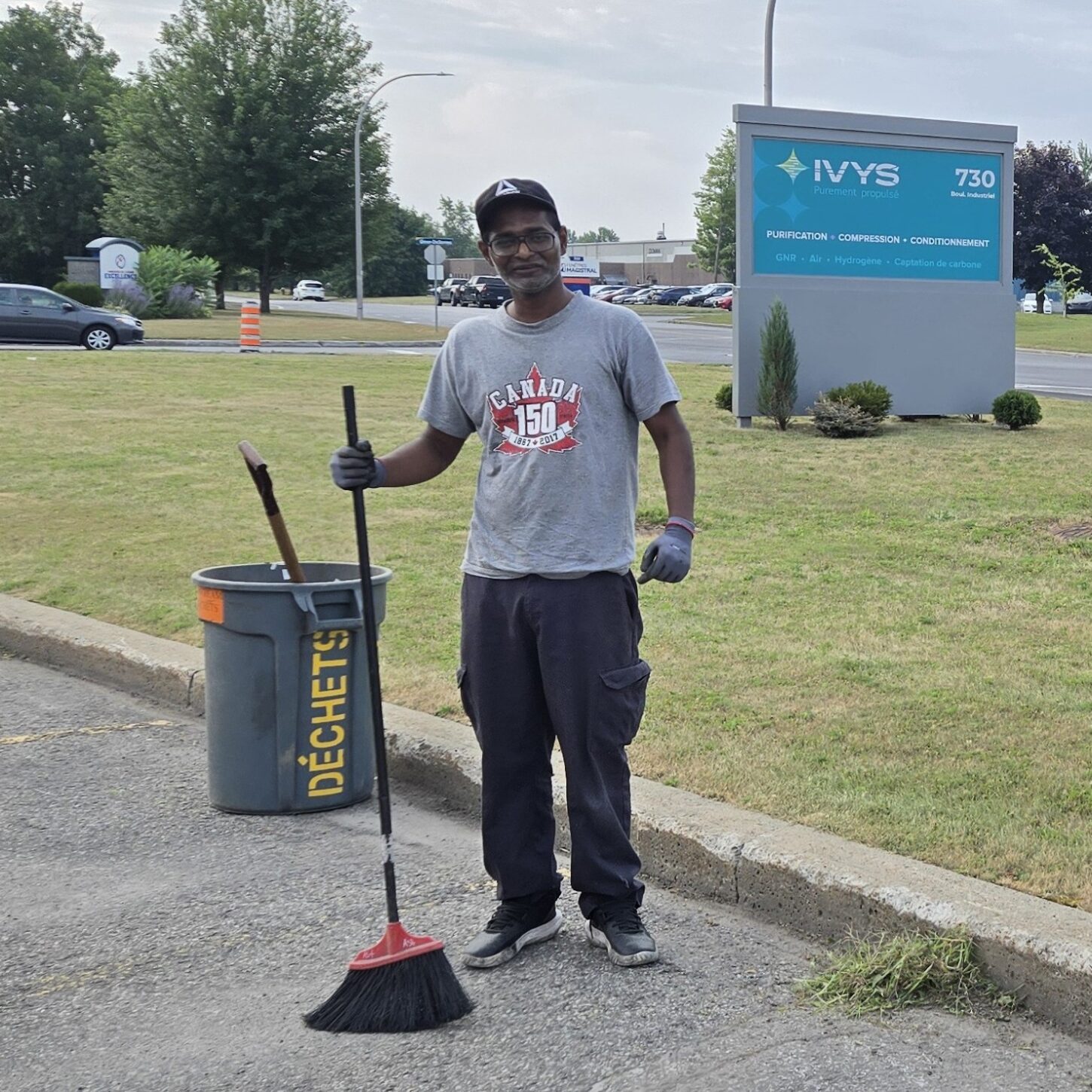 Thanushan 2 carré Thanushan, assembleur électrique chez Biostream, debout devant l’enseigne extérieure d’Ivys, tenant un balai à la main. Il s’est porté volontaire pour nettoyer le stationnement. Il a une expression de fierté.