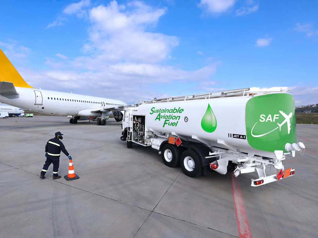 une scène sur le tarmac d’un aéroport, où un camion-citerne de carburant est stationné à proximité d’un avion. Le camion est clairement identifié comme transportant du carburant d’aviation durable, avec l’inscription « Sustainable Aviation Fuel » (SAF) bien visible sur le flanc, accompagnée d’un symbole de goutte verte, représentant l’aspect écologique du carburant. Un travailleur en tenue de sécurité — gilet haute visibilité et casque — est en train de placer ou retirer un cône de signalisation orange près du camion, ce qui indique une opération en cours ou une mesure de sécurité. À l’arrière-plan, on aperçoit un avion stationné.